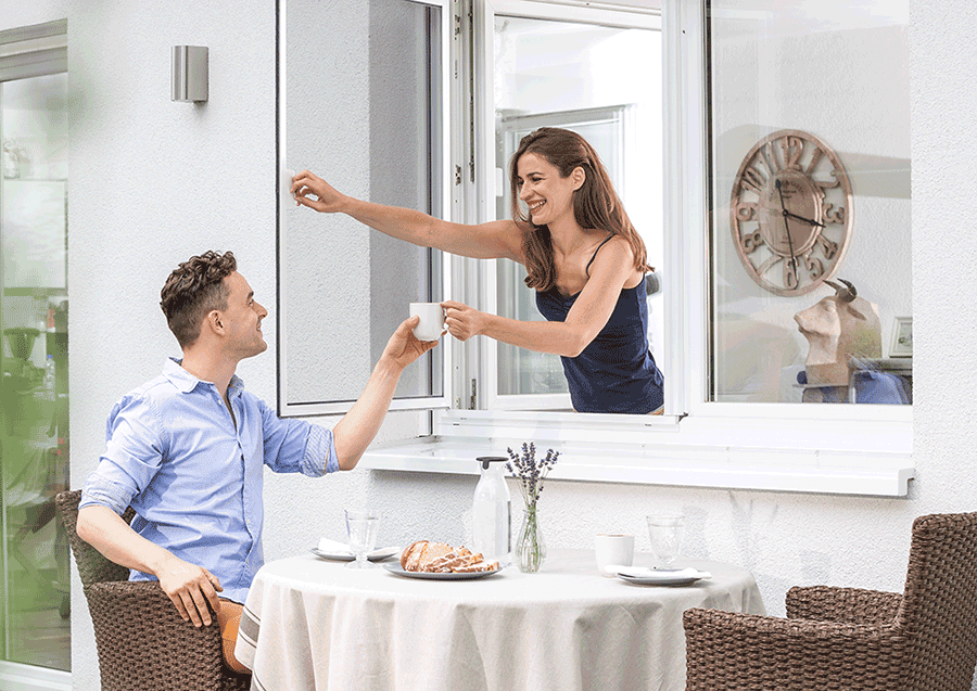 Mann ist am Frühstücken. Seine Frau reicht ihm seinen Kaffee durchs Pendelfenster.