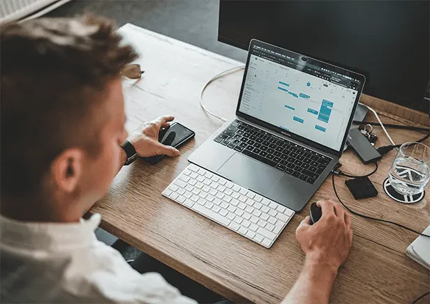 L'uomo lavora alla scrivania con un computer portatile su cui è aperto un calendario digitale; utilizza una tastiera e un mouse esterni tenendo in mano uno smartphone.